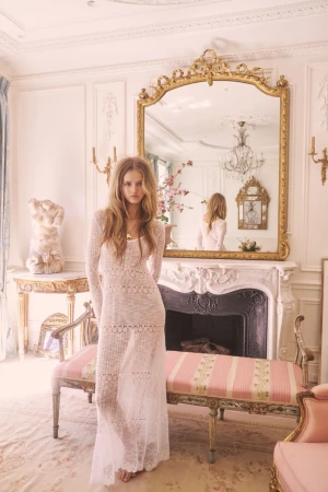 A fair-skinned young woman in a long white crochet dress stands in an ornate room with antique furniture and a decorative fireplace.