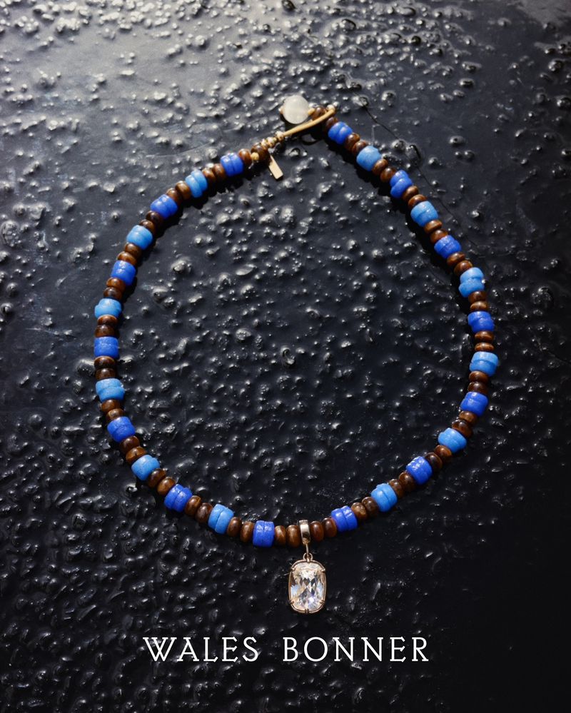 Beaded necklace featuring blue and brown tones with a hanging pendant, presented against a dark, textured background. Wales Bonner logo visible.