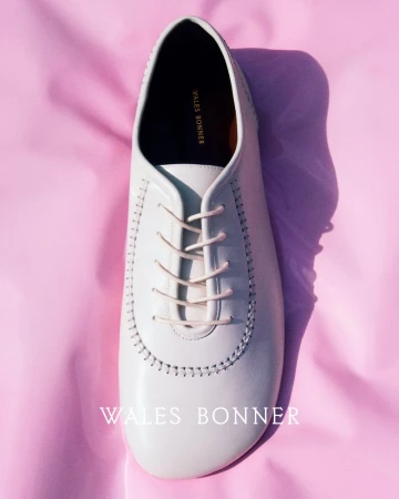 Elevated shot showcases a single, off-white leather dress shoe with matching laces, set against a soft pink fabric backdrop.
