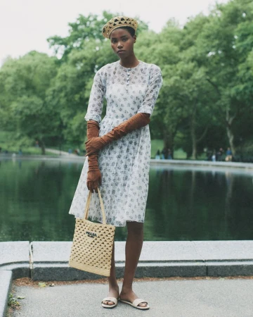 A woman poses in a floral dress, woven hat, and gloves by a pond in a park on a cloudy day, holding a woven Prada handbag.