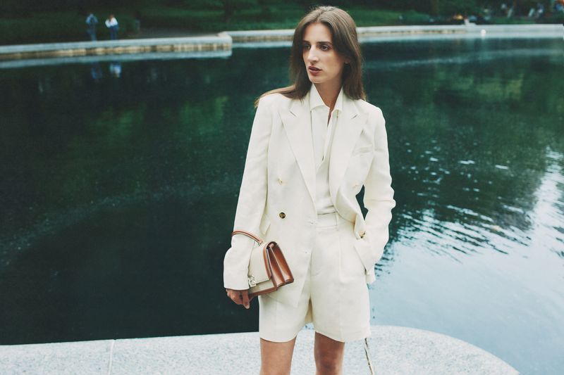Woman in a cream shorts suit and matching handbag poses in front of a lake with soft lighting and a natural outdoor setting.