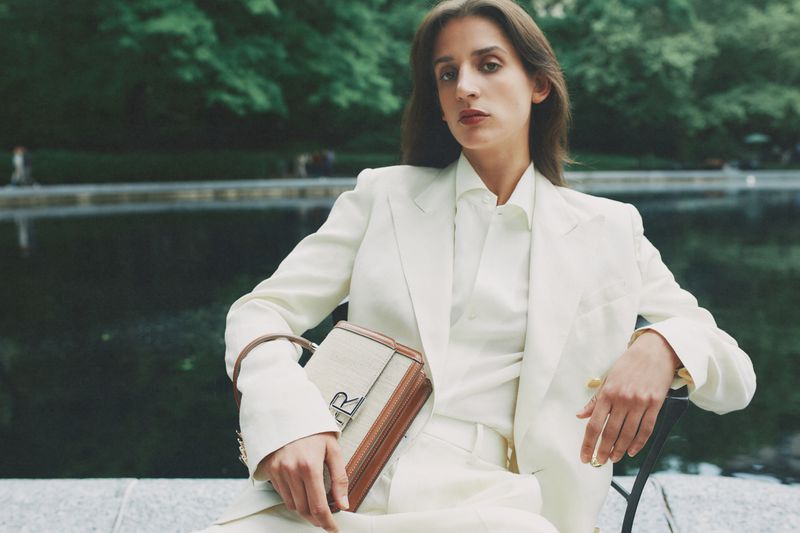 Woman in a white suit poses outdoors with a structured handbag, exuding elegance and style in a blurred park setting.