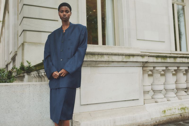 An adult female model poses in a blue suit in front of an urban stone building with a neutral expression and an overcast day-lit scene.