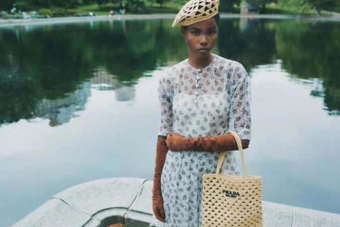 Woman poses near lake wearing floral dress, beret, and holding Prada bag. Water and foliage provide a serene backdrop.