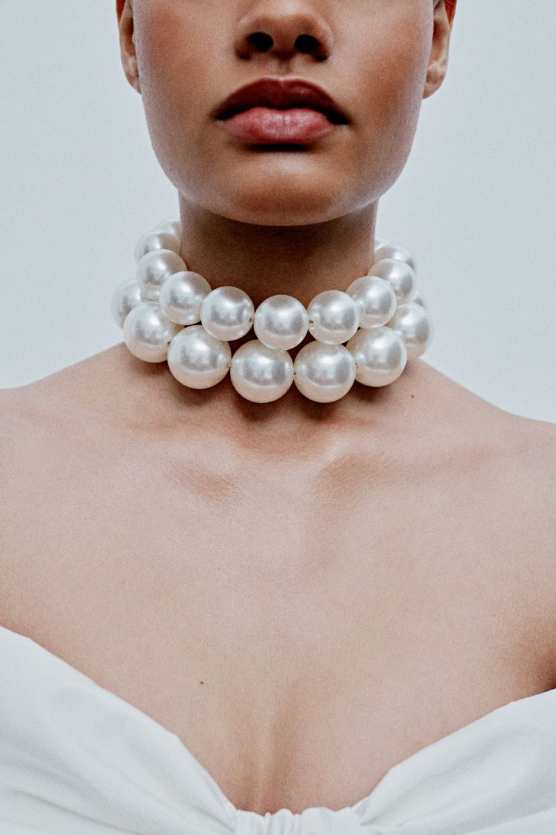 Close-up of an adult female wearing a large pearl necklace against a neutral background, showcasing fashion and beauty.