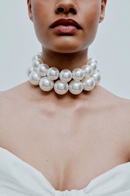 Close-up of an adult female wearing a large pearl necklace against a neutral background, showcasing fashion and beauty.
