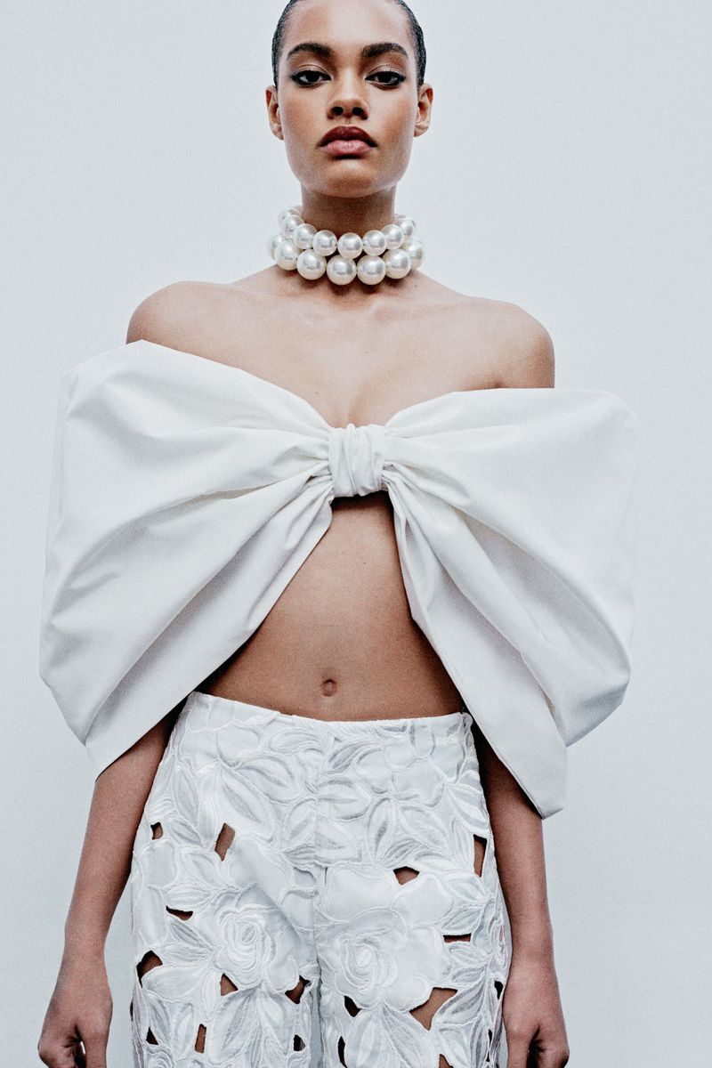 Fashion portrait of a woman wearing a white bow top and cut-out pants against a plain background, showcasing modern, elegant couture.
