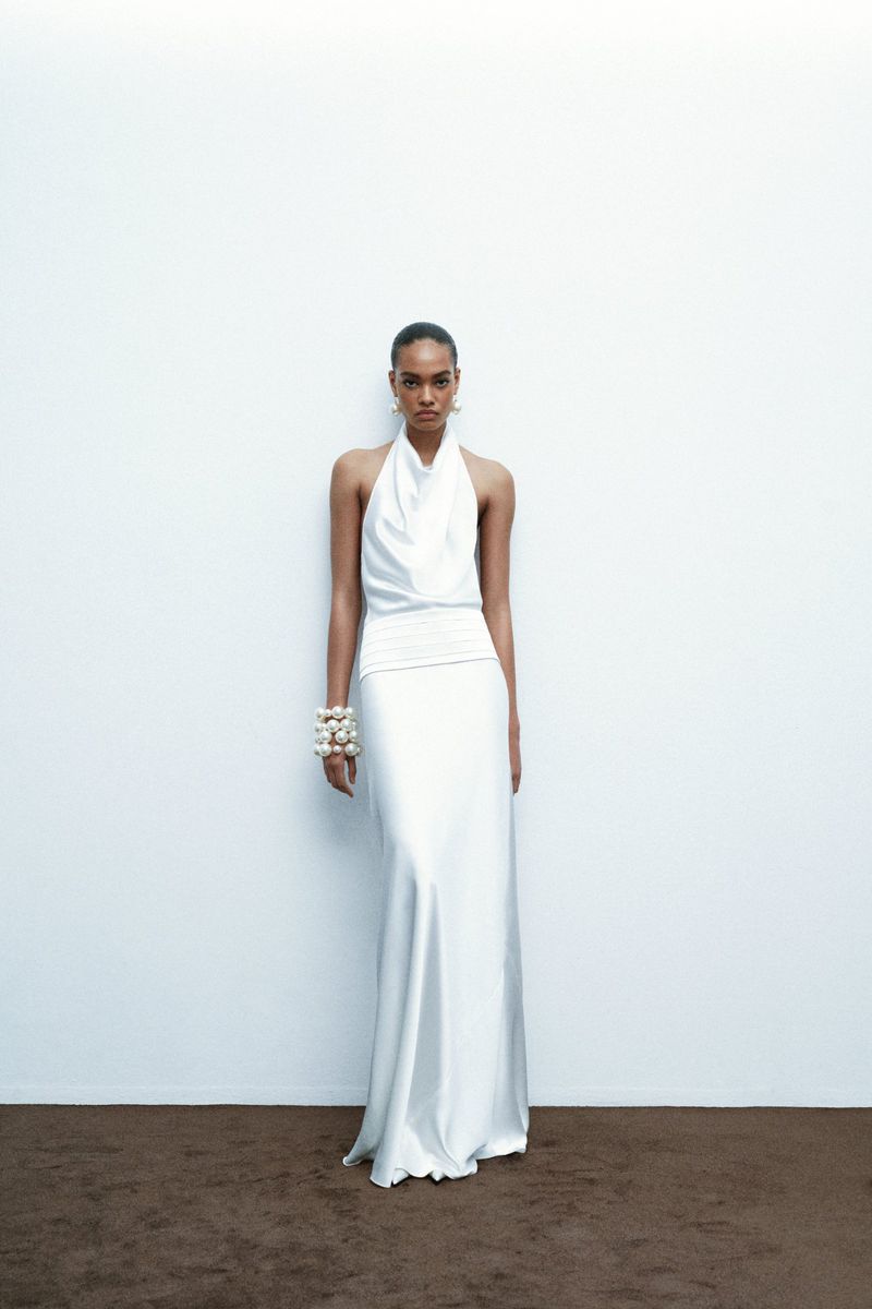 Woman in white halter gown stands against a white wall. She wears pearl bracelets and earrings. Soft, diffused lighting creates subtle shadows.