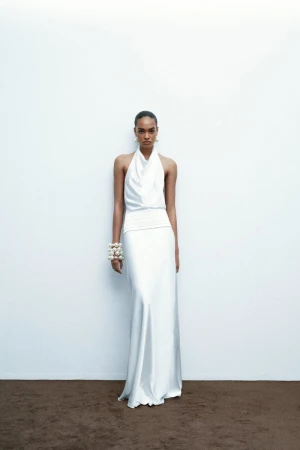 Woman in white halter gown stands against a white wall. She wears pearl bracelets and earrings. Soft, diffused lighting creates subtle shadows.