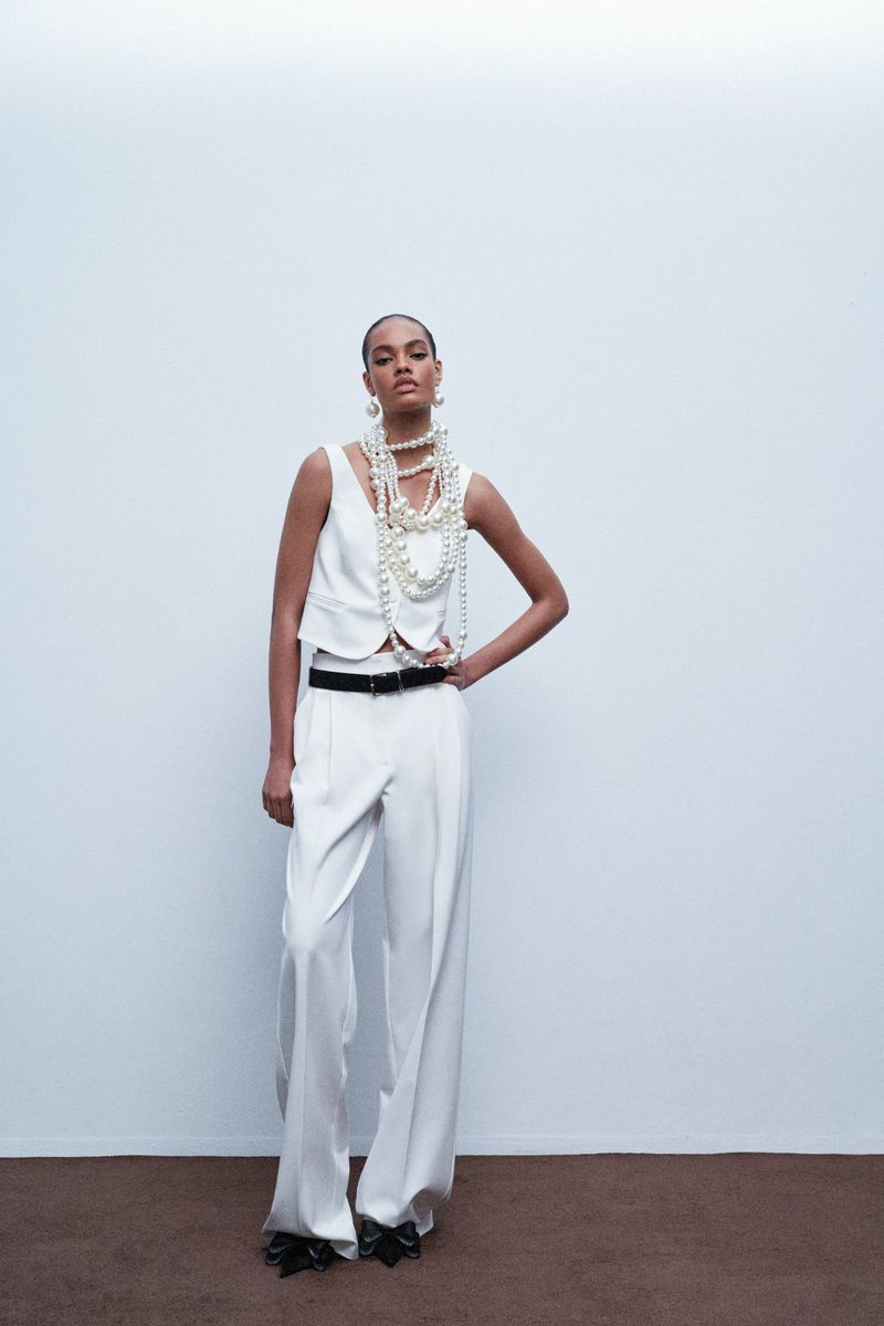 Elegant woman in a white pantsuit poses with multiple pearl necklaces against a plain background in a fashion editorial shot.