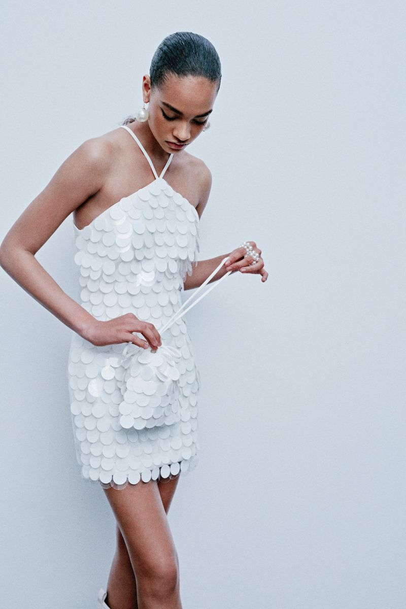 A young adult female poses in a white, sequined halter dress with a matching drawstring bag, against a white background in a studio shot.