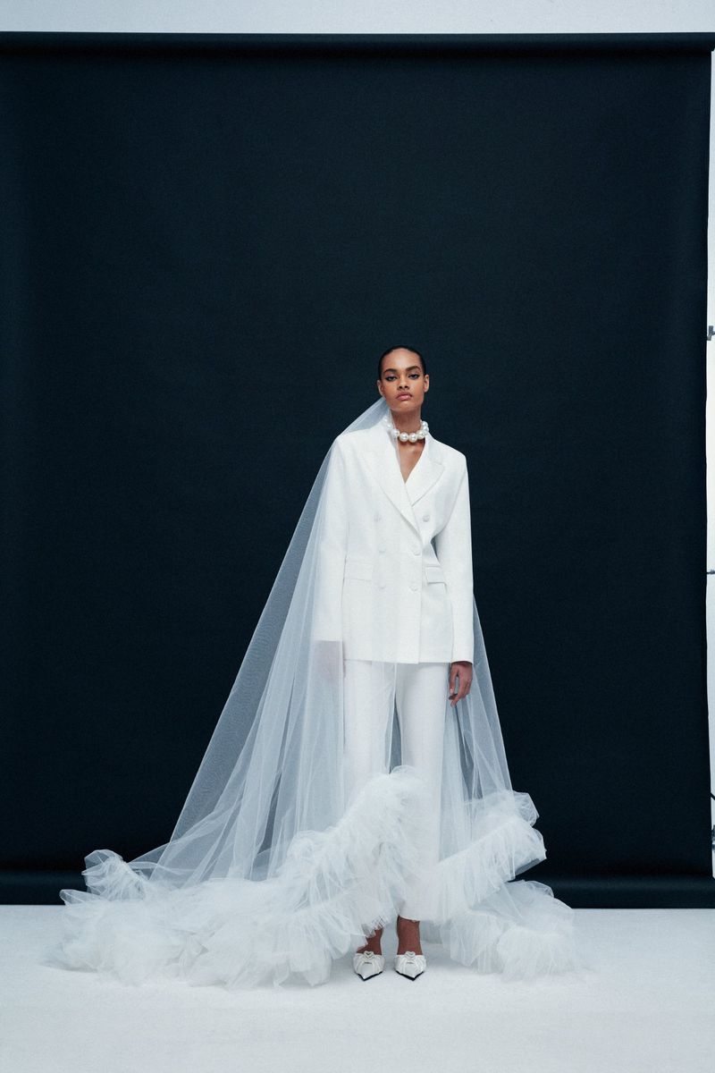 Woman in white suit with tulle ruffles and veil poses against a black backdrop in a fashion setting. She wears a pearl necklace.