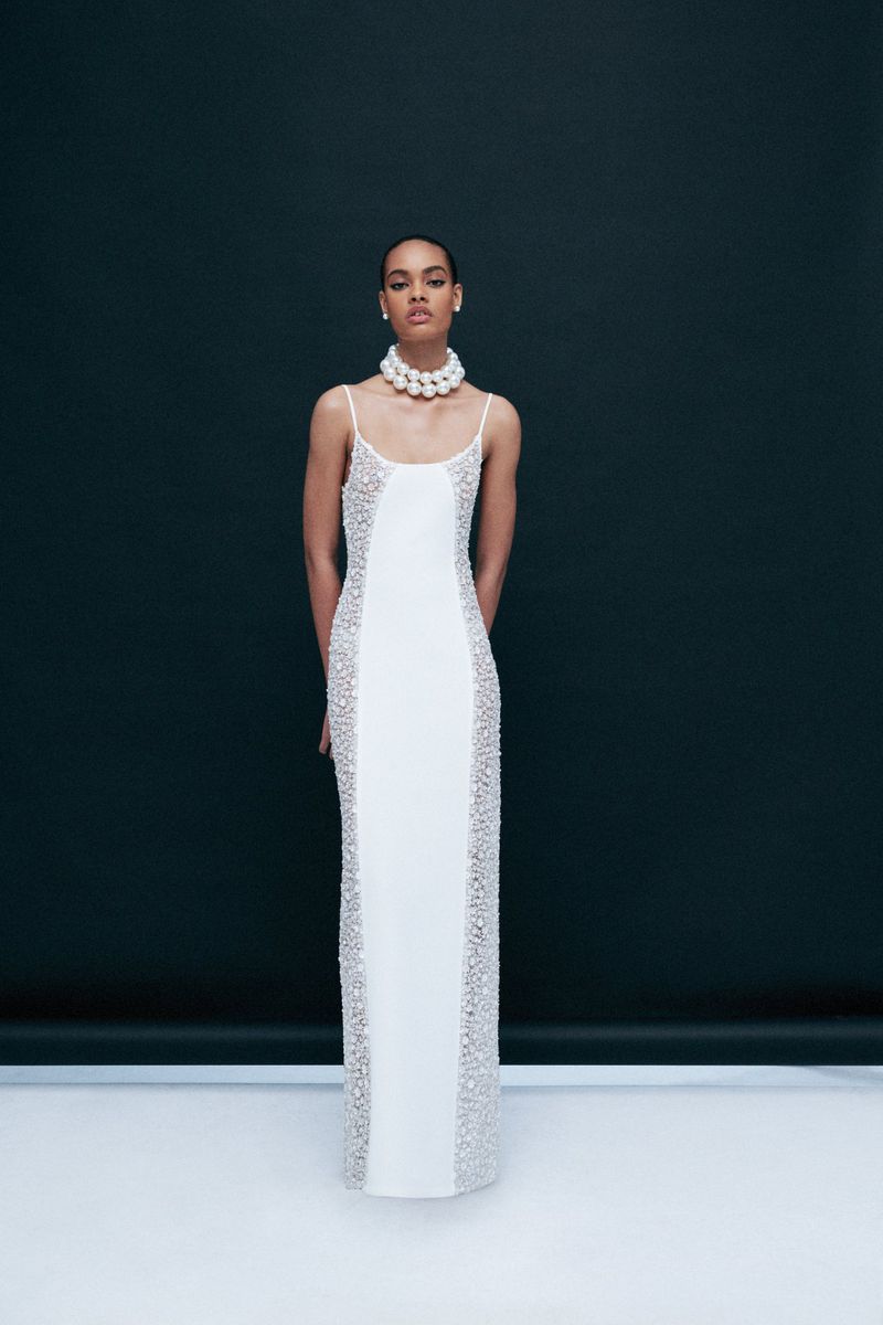 Elegant woman in a white gown with beaded details poses against a dark backdrop, accessorized with a pearl choker necklace.