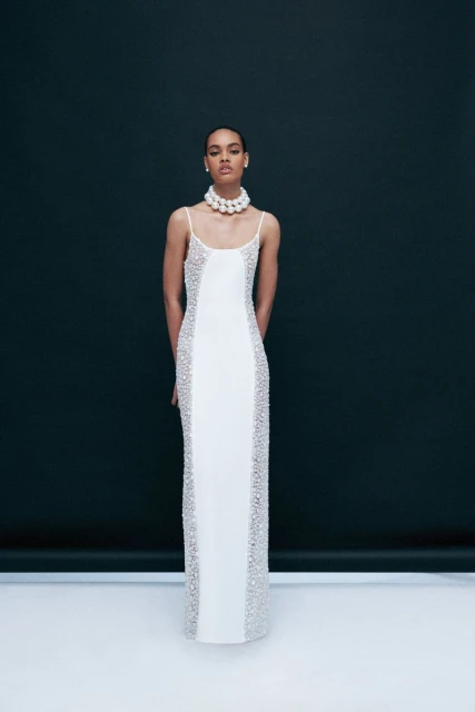 Elegant woman in a white gown with beaded details poses against a dark backdrop, accessorized with a pearl choker necklace.