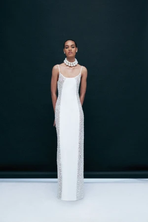Elegant woman in a white gown with beaded details poses against a dark backdrop, accessorized with a pearl choker necklace.