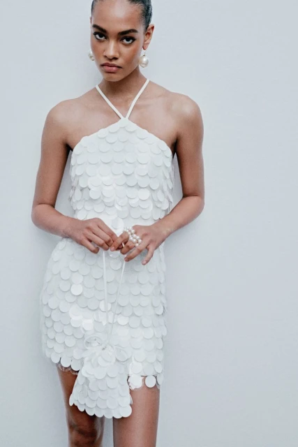 Woman poses in a short white halter dress with sequin embellishments, holding a matching drawstring bag, against a pale background.