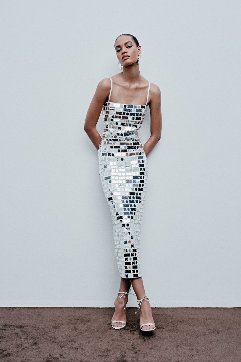 Woman posing in a reflective mosaic-tile dress in front of a plain wall with visible strappy heels and neutral studio setting.