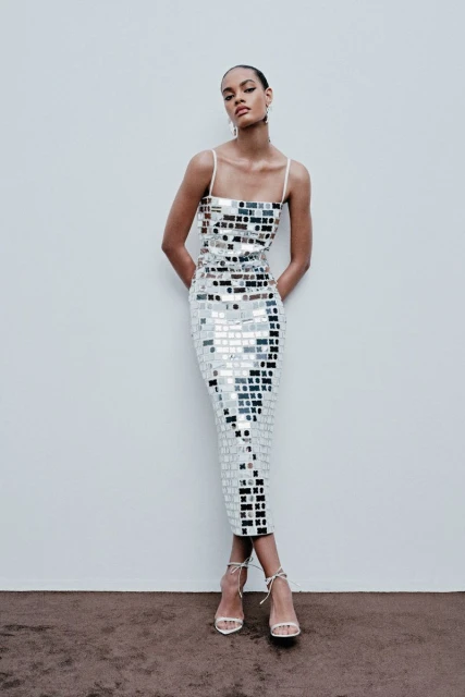 Woman posing in a reflective mosaic-tile dress in front of a plain wall with visible strappy heels and neutral studio setting.
