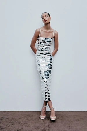 Woman posing in a reflective mosaic-tile dress in front of a plain wall with visible strappy heels and neutral studio setting.