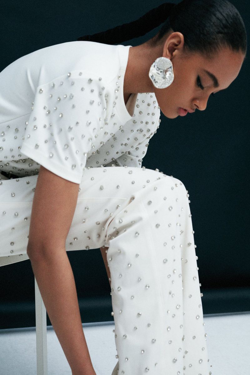 Elegant adult female with a ponytail poses in a studio wearing an off-white beaded top and matching pants and large earrings.