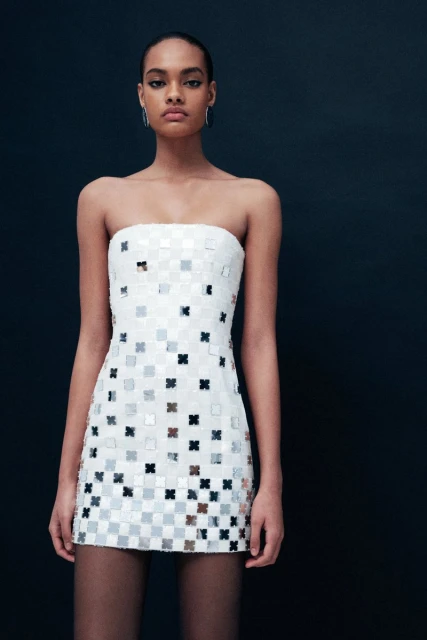Elegant woman in a white strapless mini dress with square embellishments, posing against a dark background in a fashion studio shot.