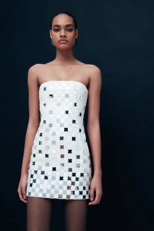 Elegant woman in a white strapless mini dress with square embellishments, posing against a dark background in a fashion studio shot.