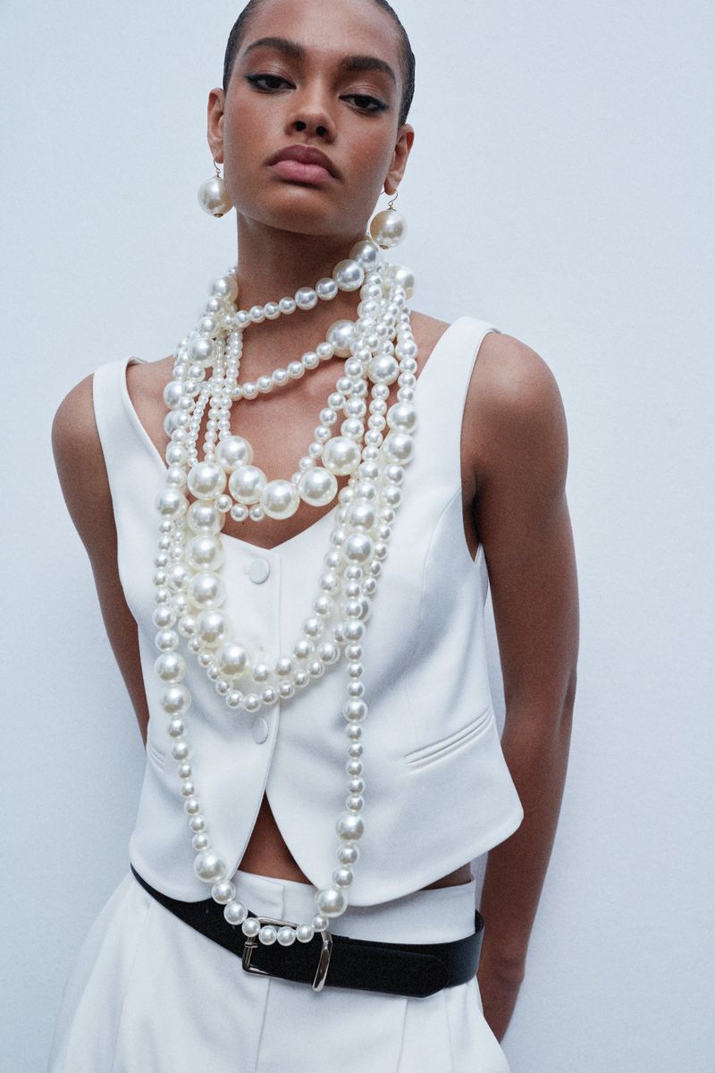 A woman in a white vest poses against a white background, adorned with layered pearl necklaces and earrings, showcasing fashion and style.