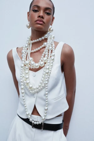 A woman in a white vest poses against a white background, adorned with layered pearl necklaces and earrings, showcasing fashion and style.