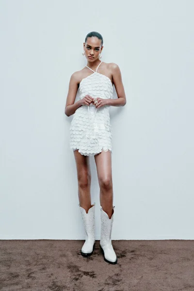 Model in a white halterneck mini dress and white cowboy boots against a plain background for ILA Bridal 24.