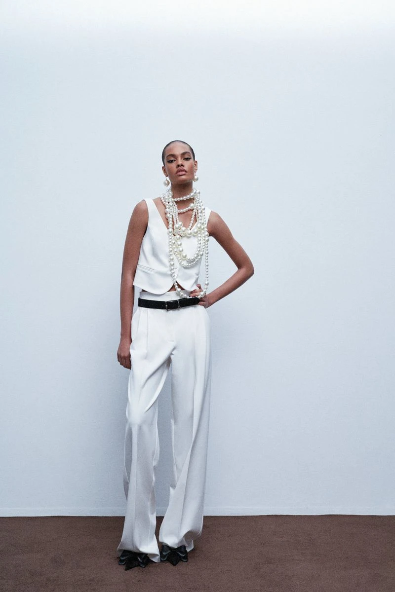 Model wearing white trousers and vest with layered pearl necklaces against white seamless background