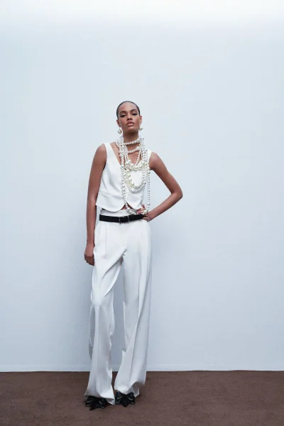 Model wearing white trousers and vest with layered pearl necklaces against white seamless background
