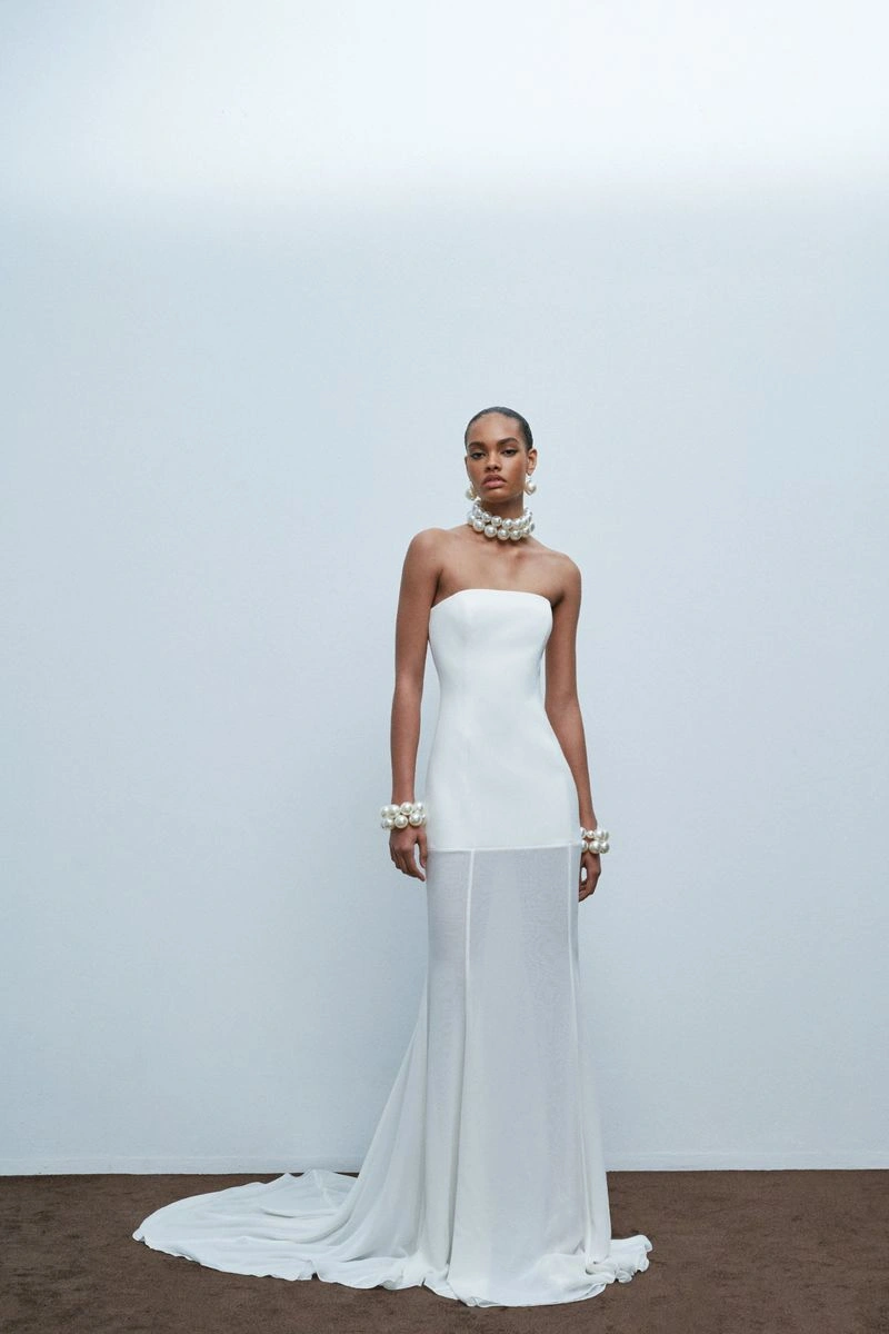 Model wearing a strapless white wedding dress and pearl accessories against a pale studio background