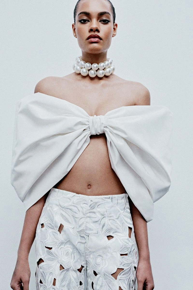 Model wearing a white strapless bow top, pearl choker, and cutout trousers against a plain white background.