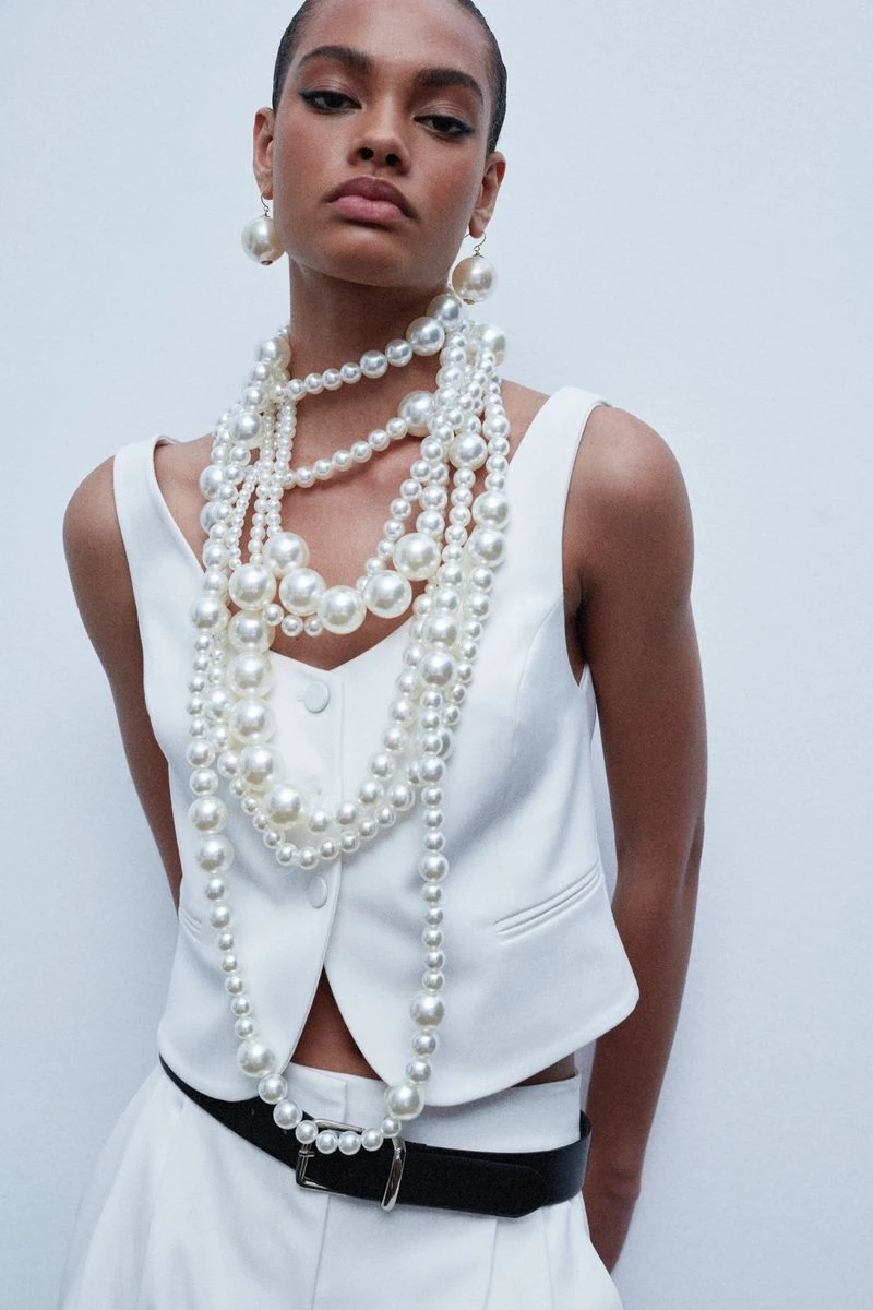 Model wearing a white vest layered with oversized pearl necklaces against a seamless white background