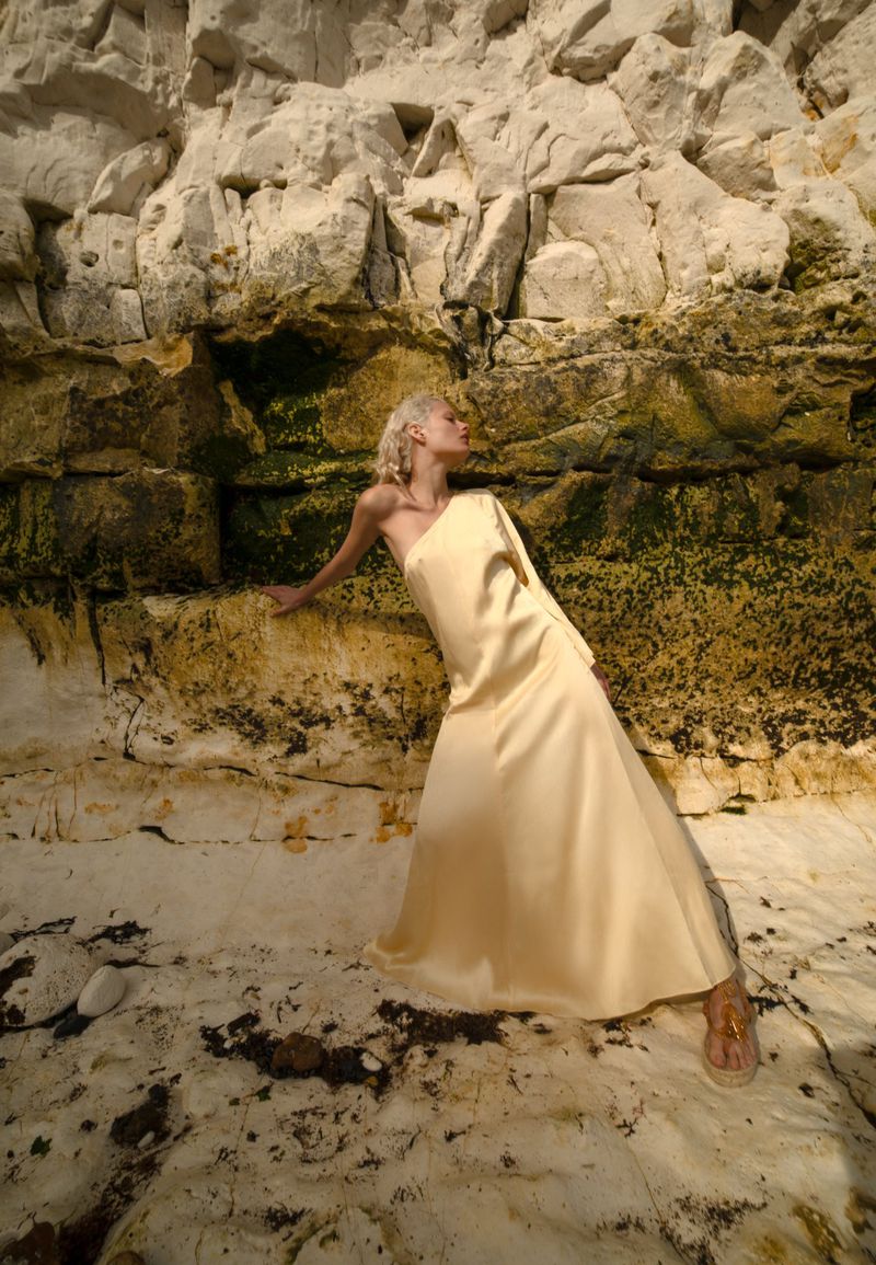 Woman in flowing yellow dress posing against weathered rock formations in outdoor setting, bathed in natural light, and elegant sandals.