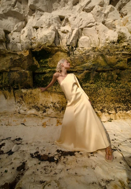 Woman in flowing yellow dress posing against weathered rock formations in outdoor setting, bathed in natural light, and elegant sandals.