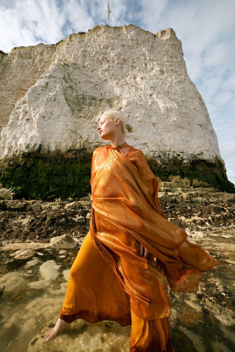 Woman in orange dress stands barefoot on a rocky shore with cliffs in the background and a bird flying overhead in a fashion shoot.