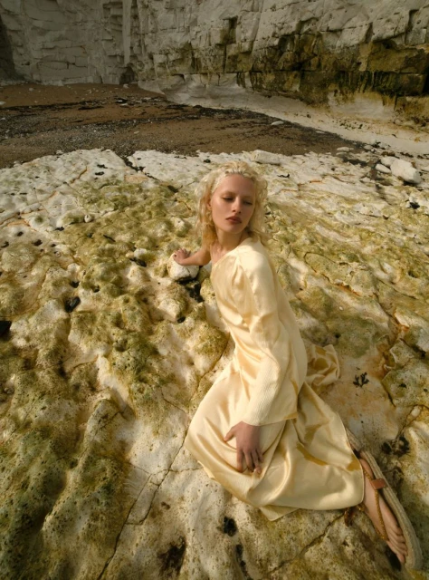 Woman in a long, pale yellow dress lying on a rocky beach near a white cliff. The model is blond with short curly hair.