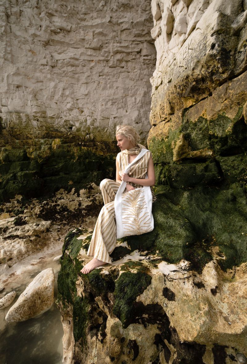 Woman in striped outfit sits on mossy rocks by a stone cliff with a white tote bag, ocean visible nearby, posing in natural light.