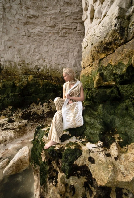 Woman in striped outfit sits on mossy rocks by a stone cliff with a white tote bag, ocean visible nearby, posing in natural light.