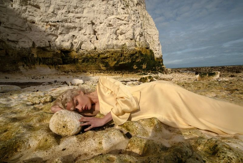 Woman in yellow dress rests on a rock at the beach by a chalk cliff under a cloudy sky, creating a dreamy, surreal outdoor scene.