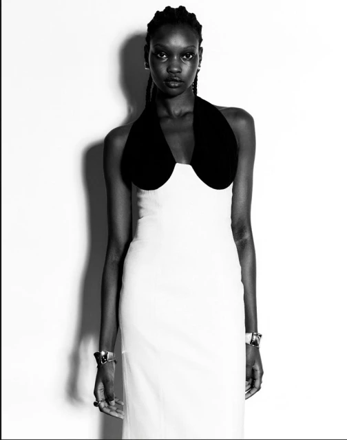Black and white studio portrait of a slender Black woman in a white dress with a black halter top, looking at the camera with a serious expression.