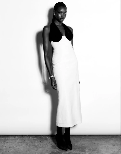 Elegant black and white fashion shot of a slender woman in a form-fitting white midi dress with black trim and black heels, against a plain background.