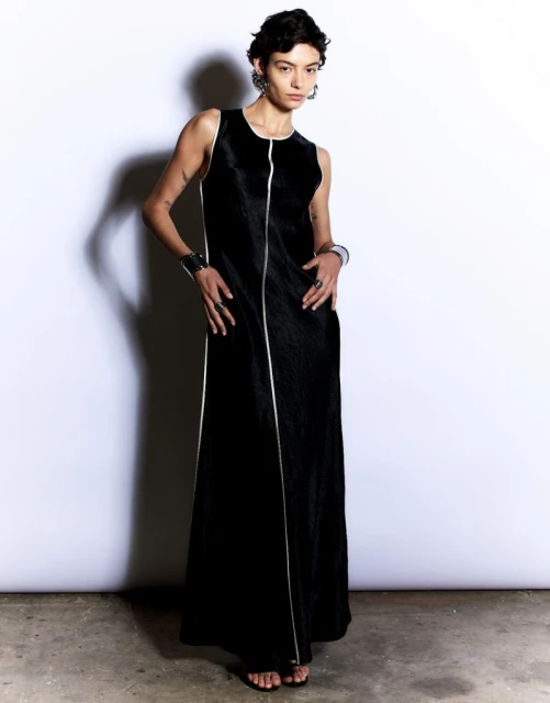 A model poses in a full-length black sleeveless dress with white trim, wearing hoop earrings and metal cuffs, against a white background.