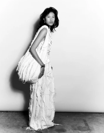 A stylish young woman in a textured white dress poses in a studio, set against a bright white backdrop with natural lighting.