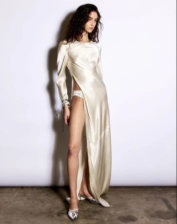 A slender fashion model in a shimmering white dress with a thigh-high slit poses in a photo studio with a minimalist white background.