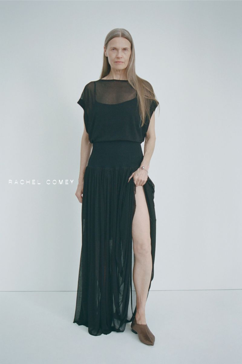 Full-length portrait of a mature adult female in a black dress with a thigh-high slit, standing against a white background in a studio.