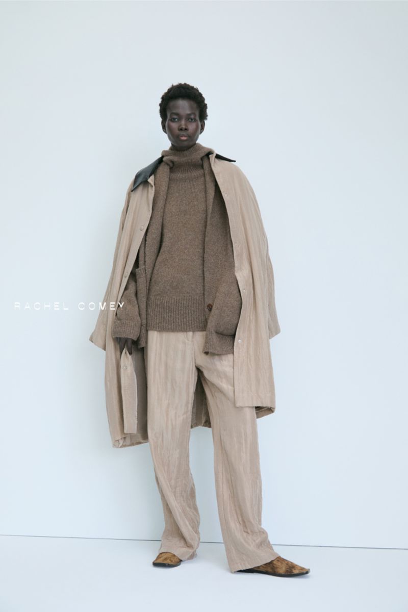 A dark-skinned model poses in neutral-tone separates: a brown sweater, trousers, and a beige trench coat against a stark white background.