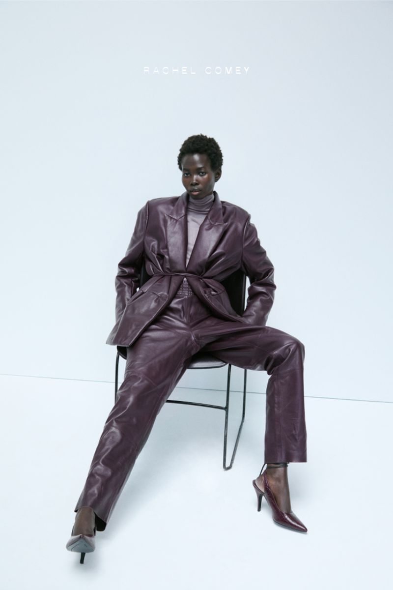 An adult female fashion model in a plum-colored leather suit and heels is seated in a studio with a light backdrop.