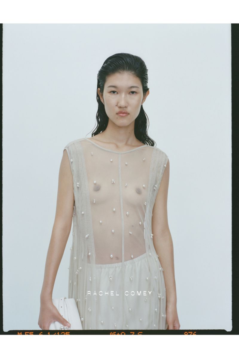 Portrait of an Asian female fashion model wearing a sheer beige dress with bead accents, holding a white clutch against a bright backdrop.